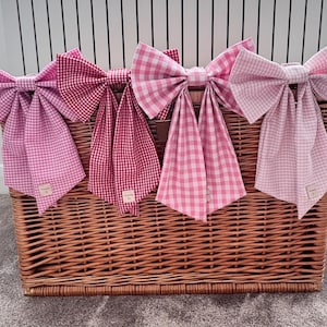 May include: Four large fabric bows with long tails, each featuring a different shade of pink and red gingham check pattern. The bows are arranged in a row, displaying variations from light pastel pink to a deeper, vibrant red checkered design.