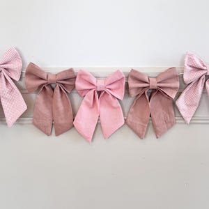 May include: A horizontal garland featuring five fabric bows strung together. The two outer bows have a light pink and white gingham pattern, while the three inner bows are solid shades of dusty rose, soft pink, and a slightly darker mauve pink.