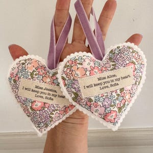 May include: Two heart-shaped fabric ornaments with a floral pattern in pink, purple, and blue. Each features a white scalloped edge, a lavender velvet ribbon loop, and a beige fabric label printed with the text: Miss Alice, I will keep you in my heart. Love, Sofia, or Miss Jessica, I will keep you in my heart. Love, Sofia.