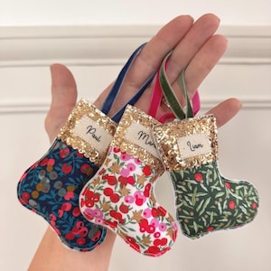 May include: Three small fabric stockings with gold glitter cuffs, each featuring a unique floral pattern in navy, white, and green. Each stocking has a white fabric label with the embroidered names Paul, Maria, and Liam, and a velvet ribbon loop.