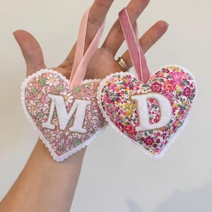 May include: Two heart-shaped fabric ornaments featuring floral patterns in shades of pink, white, and green. Each heart is edged with white scalloped trim, includes a pink velvet ribbon loop, and displays a large white letter M or D.