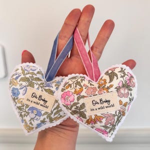 May include: Two heart-shaped fabric ornaments with floral patterns in blue, pink, and green. Each heart features a white scalloped edge and a beige fabric label printed with the text Oh Baby its a wild world. One has a blue velvet ribbon, the other a pink.