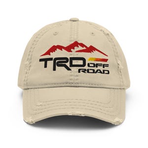 May include: A distressed beige baseball cap featuring the embroidered text "TRD OFF ROAD" in black, red, and yellow. The design includes a mountain range graphic above the text, and the cap has a worn, vintage look.