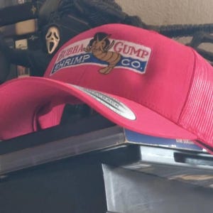 以下が含まれることがあります： 明るいピンク色のメッシュバックとカーブしたつばのトラッカーハット。帽子の前面には「Bubba Gump Shrimp Co.」の文字と漫画のエビが描かれたパッチが付いています。この帽子はおそらくお土産または商品アイテムです。