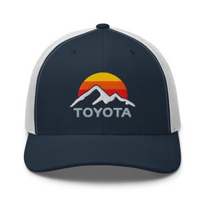 May include: Navy blue and white trucker hat with an embroidered mountain and sun design, and the word "TOYOTA" in white. The hat has a curved peak and a mesh back for ventilation.