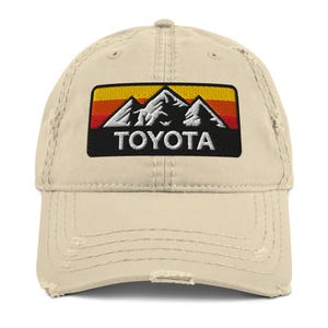May include: A distressed beige baseball cap with a rectangular patch. The patch features a mountain range design in white, orange, and black, and the word "TOYOTA" in white block letters.