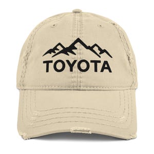 May include: A beige baseball cap with a distressed finish, displaying a black mountain range graphic and the word "TOYOTA". The cap has a worn appearance with frayed edges and a curved peak. It's a classic six-panel design.