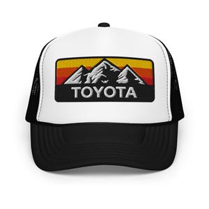 May include: A black and white trucker hat with a white front panel. The hat features an embroidered patch with a mountain range design in white, orange, and red, with the word "TOYOTA" in white block letters.