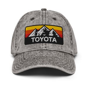 May include: A grey, vintage-style baseball cap with a mountain and sunset design. The embroidered patch features white mountains, a yellow, orange, and red gradient, and the word "TOYOTA" in white.