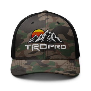 May include: Camouflage trucker hat with a black mesh back and a curved brim. The front features an embroidered mountain range design with a rising sun and the text "TRDPRO" in white.