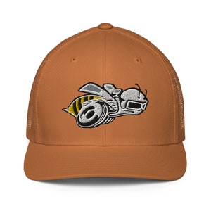 Puede incluir: Gorra de camionero naranja quemado con un logotipo de abeja bordado en blanco y amarillo. La gorra tiene una parte trasera de malla y una visera curva. El diseño de la abeja es estilizado y detallado, con un casco y elementos mecánicos.