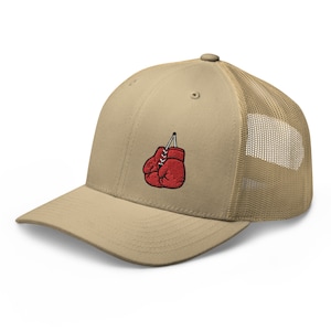 Könnte beinhalten: Beige Trucker-Cap mit rotem, gesticktem Boxhandschuh-Design. Die Kappe hat einen gebogenen Schirm, eine Mesh-Rückseite und einen Schnappverschluss. Der Boxhandschuh ist mit weißen Schnürsenkeln und einer schwarzen Aufhängeschnur versehen.
