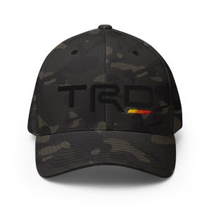 May include: A black camouflage baseball cap with the embroidered letters "TRD" in black. The "D" is accented with a horizontal stripe of yellow, orange, and red. The cap has a curved brim.