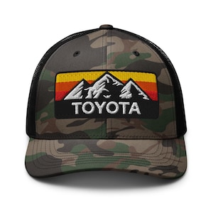 Könnte beinhalten: Tarnfarbene Trucker-Kappe mit schwarzem Mesh-Rücken und gebogenem Schirm. Auf der Vorderseite befindet sich ein rechteckiger Aufnäher mit einem Bergketten-Design in Weiß, Orange, Gelb und Rot, mit dem Wort "TOYOTA" in Weiß.