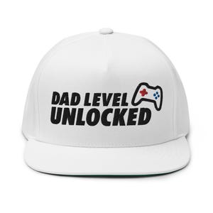 May include: A white flat-brimmed hat with the text "DAD LEVEL UNLOCKED" in bold black letters. A controller graphic with red and blue accents is to the right of the text. The hat has a green underside.