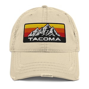 May include: A beige distressed baseball cap with an embroidered patch featuring a mountain range and the word "TACOMA" in black text.