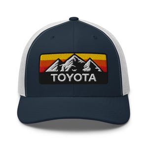 May include: A navy blue and white trucker hat with a mountain range logo. The logo features a white mountain range with an orange and red sunset gradient. The word "TOYOTA" is embroidered in white.