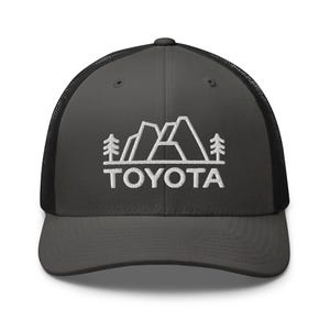 May include: A dark grey trucker cap with a black mesh back. The front displays a white embroidered mountain and tree design, with the word "TOYOTA" stitched beneath it.