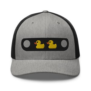 May include: A gray trucker hat with a black mesh back and a black rectangular patch. The patch features two embroidered yellow rubber duckies with orange beaks.