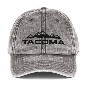 May include: A gray distressed denim baseball cap with black embroidered mountains and the word "TACOMA" underneath.