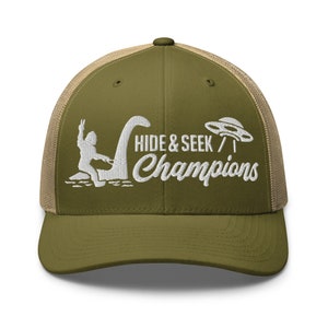 May include: Olive green and tan trucker hat with the embroidered text "HIDE & SEEK / Champions". The design includes a white silhouette of the Loch Ness Monster, a UFO, and a figure giving a peace sign.