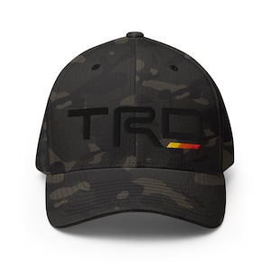 May include: A black camouflage baseball cap featuring the letters TRD embroidered in black on the front. The letter D includes a small horizontal stripe with yellow, orange, and red gradient colors positioned underneath the right side.