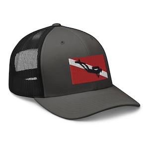 May include: A charcoal gray trucker hat with a black mesh back. The hat features an embroidered patch with a red and white dive flag and a black diver silhouette. The hat has a curved brim and a snapback closure.