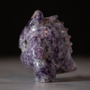 Pu&ograve; includere: Una piccola scultura di drago in ametista intagliata di colore viola. Il drago ha una schiena spinosa e una lunga coda.
