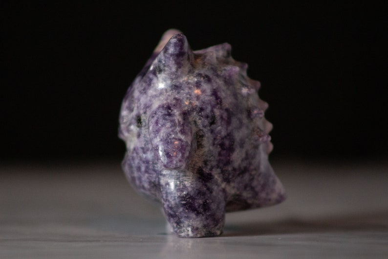 Pu&ograve; includere: Una piccola scultura di drago in ametista intagliata di colore viola. Il drago ha una schiena spinosa e una lunga coda.