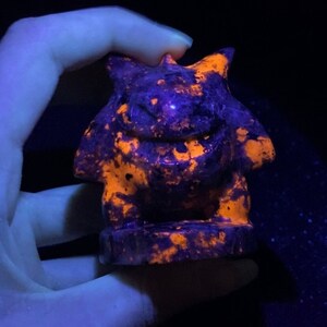 Pu&ograve; includere: Una piccola statuetta scolpita di una creatura mitologica, forse un drago o un grifone, realizzata in pietra di colore viola scuro e arancione. La statuetta brilla sotto una luce nera, rivelando i colori arancione e viola.