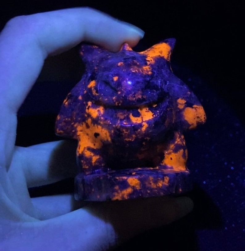 Pu&ograve; includere: Una piccola statuetta scolpita di una creatura mitologica, forse un drago o un grifone, realizzata in pietra di colore viola scuro e arancione. La statuetta brilla sotto una luce nera, rivelando i colori arancione e viola.