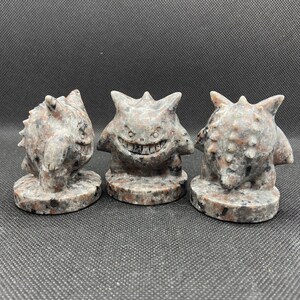 Pu&ograve; includere: Tre piccole figurine di pietra scolpite a mano di un personaggio Pokemon. Le figurine sono grigie con macchie nere e hanno un'espressione minacciosa.