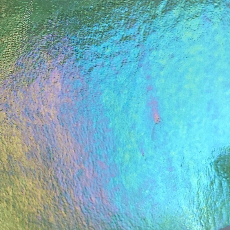 May include: Abstract close-up of a shimmering, iridescent surface. The colors shift from green to blue and purple, creating a textured, oil slick effect. The surface appears to be slightly rippled, adding depth.