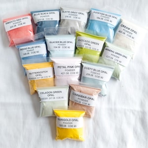 May include: Assortment of small bags containing various colored opal powders. Each bag is labeled with the color name, "POWDER", and the COE number. The colors include: Pimento Red, Blue Black, Slate Gray, Egyptian Blue, Cream, Indigo Blue, Glacier Blue, Avocado Green, Mineral Green, Butterscotch, Petal Pink, Dusty Blue, Celadon Green, Cinnabar, and Marigold.