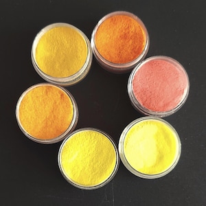 May include: A top-down view of six small, clear plastic containers filled with colorful powders. The powders are in shades of yellow, orange, and pink, arranged in a circular pattern against a dark background.