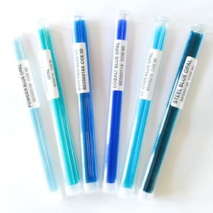 May include: Six glass rods in shades of blue, from light to dark, are displayed in clear tubes. Each tube has a white label with the text "POWDER BLUE OPAL", "EGYPTIAN BLUE OP", "COBALT BLUE OPAL", "LT. CYAN BLUE OPAL", and "STEEL BLUE OPAL".