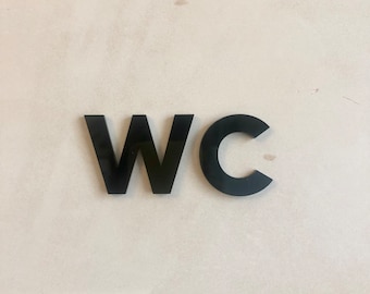 Può includere: Segno WC in acrilico nero montato su una parete beige chiaro. Le lettere sono in grassetto e senza grazie, offrendo un'estetica chiara e moderna. Il segno è progettato per i bagni pubblici.