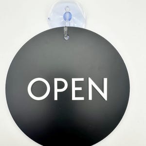 May include: A black, circular sign with the word "OPEN" in white capital letters. The sign is attached to a clear suction cup with a blue center.
