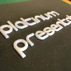 May include: White 3D letters spelling "Platinum Presented" are displayed on a dark surface. The letters are thick and appear to be made of a solid, opaque material. The letters are evenly spaced and well-lit, creating a clean and modern aesthetic.