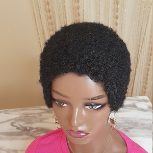 Puede incluir: Una peluca afro corta de color negro hecha de cabello sintético. La peluca está peinada en un estilo afro recortado.