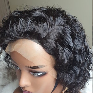 May include: A black curly human hair wig with a lace front. The wig is styled in a short bob with loose curls. The wig is on a mannequin head with a gold necklace.