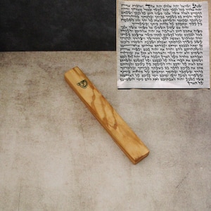 Puede incluir: Una caja de mezuzah de madera con un emblema decorativo. La caja es rectangular con un acabado natural de vetas de madera. Un pergamino con texto hebreo es visible en el fondo.