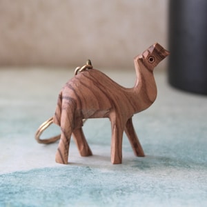 May include: A wooden camel keychain with a brass ring. The camel is carved from olive wood and has a natural wood grain.