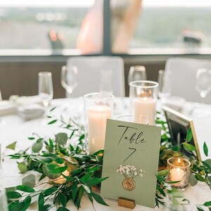 May include: A green table number card with white flowers and the text "Table 7" on a white tablecloth with greenery and candles.
