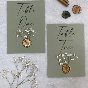 May include: Two green cards with the text "Table One" and "Table Two" written in black calligraphy. Each card has a small bouquet of white baby's breath and a gold wax seal with a leaf design.