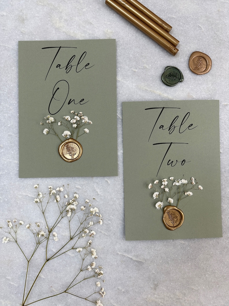 May include: Two green cards with the text "Table One" and "Table Two" written in black calligraphy. Each card has a small bouquet of white baby's breath and a gold wax seal with a leaf design.