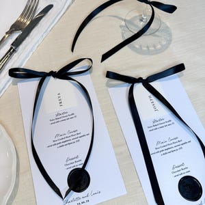 May include: Elegant wedding menu cards with black ribbon accents. Each card features a menu with starter, main course, and dessert options, sealed with a black wax stamp. A crystal wine glass and silverware are also visible.