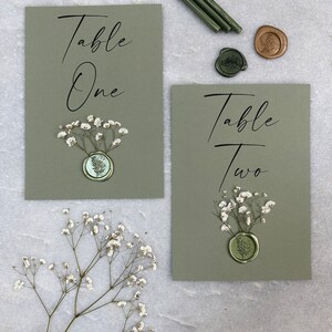 May include: Two green table number cards with white flowers and wax seals. The cards say "Table One" and "Table Two".