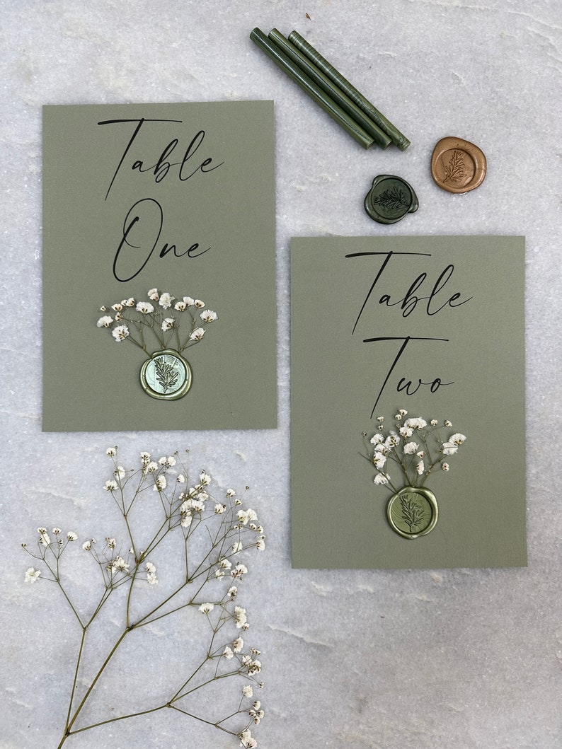 May include: Two green table number cards with white flowers and wax seals. The cards say "Table One" and "Table Two".
