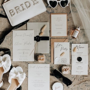 May include: A flat lay of wedding accessories including a white clutch with "BRIDE" in pearl lettering, heart-shaped sunglasses, invitations, a ring box, and white bow sandals. The invitations feature elegant calligraphy and a wax seal.